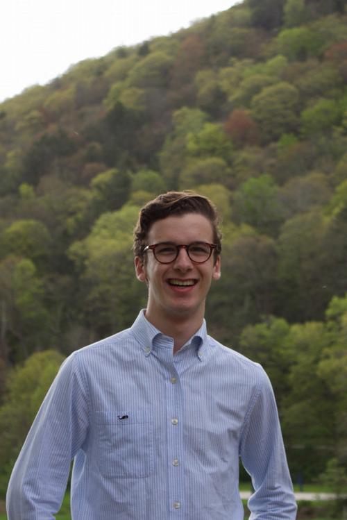 photo of Brandon Shuster smiling, wearing a light blue button down shirt, with a green forested hillside in the background